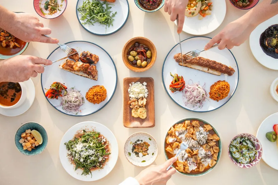 Vista cenital de una mesa de restaurante con varios platos para compartir —pan tostado, croquetas, ensaladas, aceitunas, sopa, brocheta y guarniciones— y manos de comensales sirviéndose