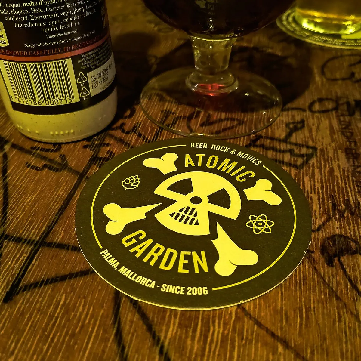 Posavasos de Atomic Garden con su logo de calavera y átomo, el lema Beer, Rock and Movies y la leyenda Palma, Mallorca desde 2006, sobre una mesa de madera junto a una cerveza