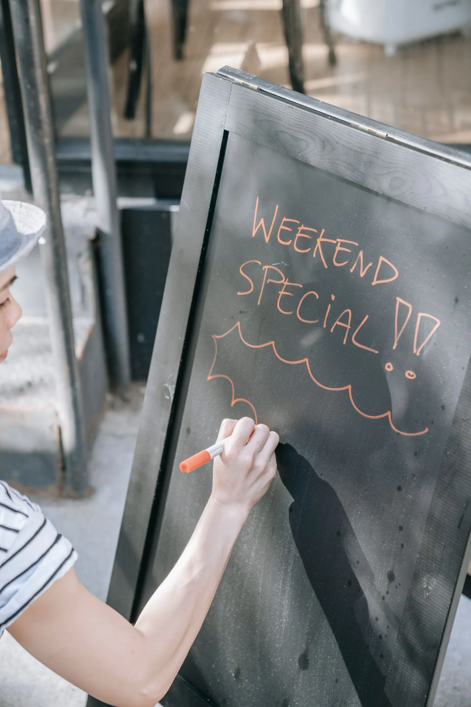 Persona escribiendo a mano el especial del fin de semana con tiza naranja en una pizarra de caballete a la entrada de un restaurante