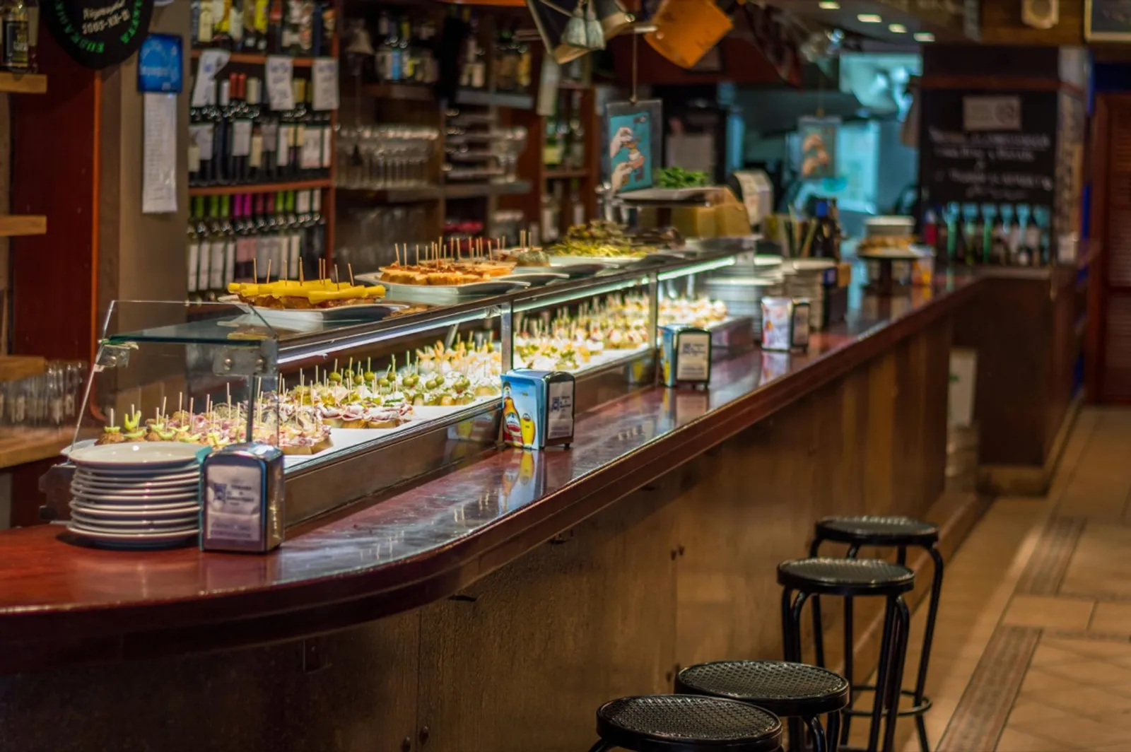Barra de bar de tapas vista en diagonal, con vitrina llena de pinchos y montaditos sobre rebanadas de pan, taburetes negros al pie de la barra y una estantería de botellas de vino al fondo